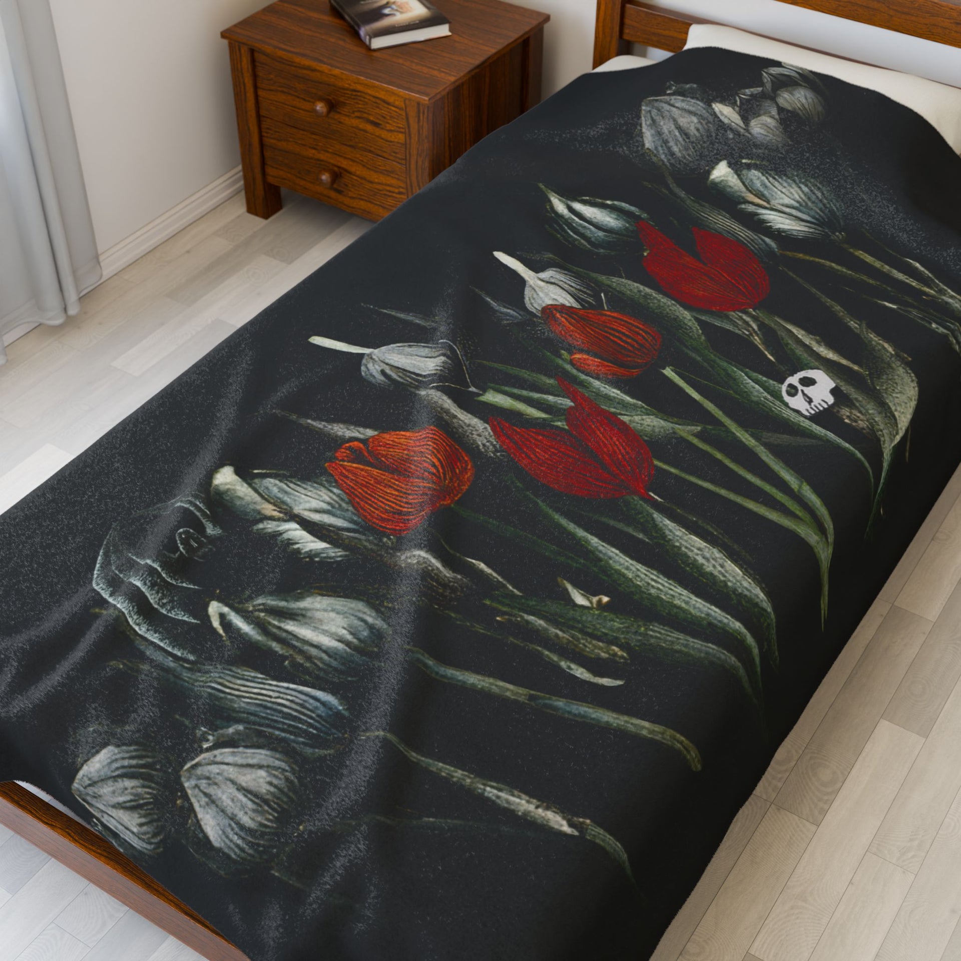 The room exudes gothic elegance with Printifys Gothic Floral Velveteen Plush Blanket in black, featuring white and red tulips and a skull motif. A wooden nightstand nearby holds cherished books, perfecting the dark yet refined atmosphere.