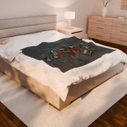 A neatly made bed showcases the Gothic Floral Velveteen Plush Blanket – Dark Grey by Printify, adorned with skulls and red roses. The room boasts gothic elegance with a modern wooden nightstand, lit lamp, matching dresser, and a light-colored rug on the wooden floor under the bed.