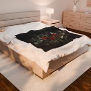 The bedroom showcases a large bed with the Gothic Floral Velveteen Plush Blanket by Printify and velveteen pillows. A wooden nightstand with a lamp stands beside it, while a classic dresser and soft rug on the wooden floor add to the rooms cozy gothic elegance.