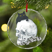The Black Cat Skull Halloween Holiday Ornament by Printify features a crystal glass design with a black cat perched on a detailed human skull, surrounded by blurred warm yellow lights, creating a perfect blend of Halloween and holiday decor for your Christmas tree.