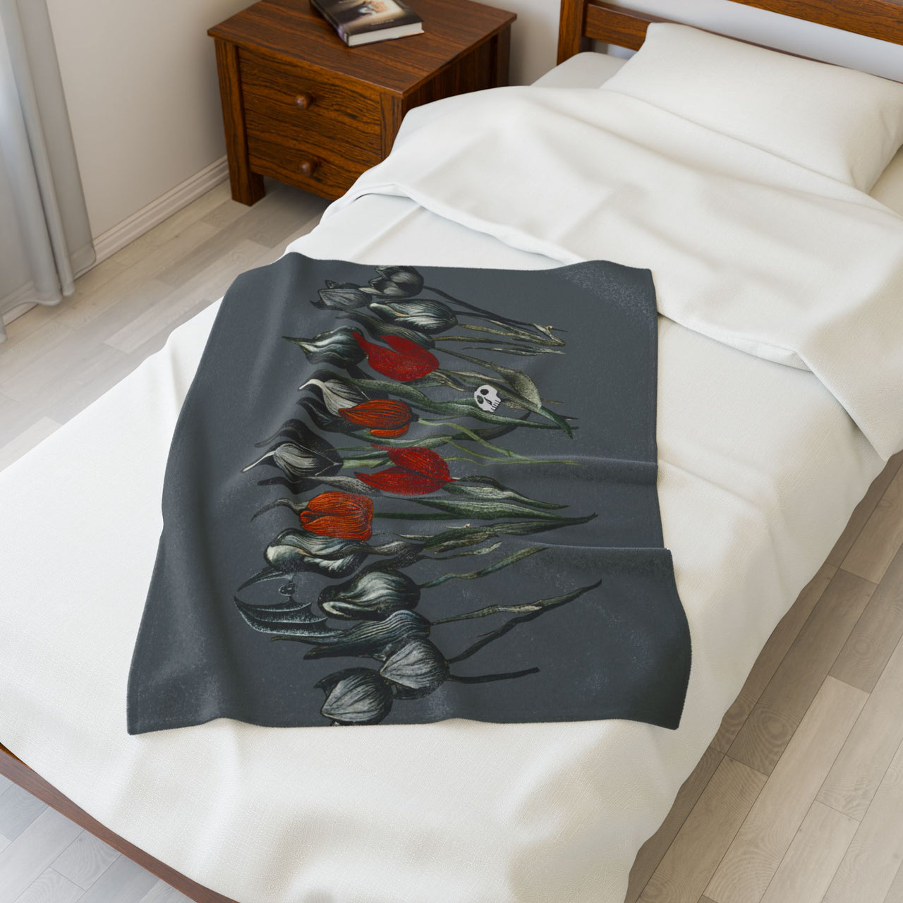 A cozy bedroom features a wooden nightstand and a bed adorned with Printifys Gothic Floral Velveteen Plush Blanket in dark grey, showcasing black and red tulips. Light-colored flooring and walls enhance the rooms gothic elegance with a warm atmosphere.