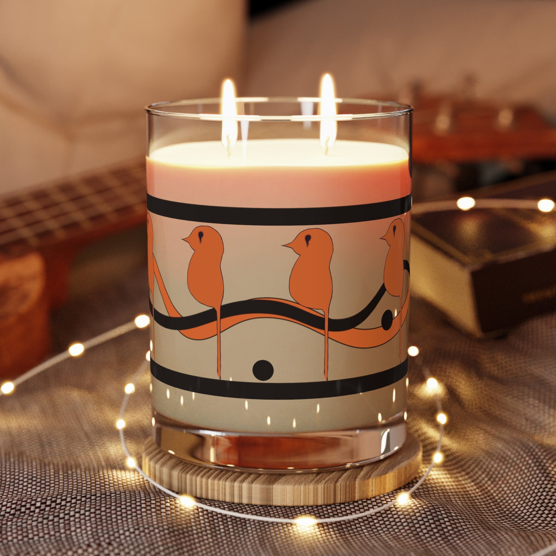 The Dream Bird Candle by Printify features a bird illustration on a soy wax candle in a glass holder, resting on a wooden coaster with warm string lights around it. A blurred ukulele is in the background. Enjoy its unique blood orange scent and long 65-hour burn time.