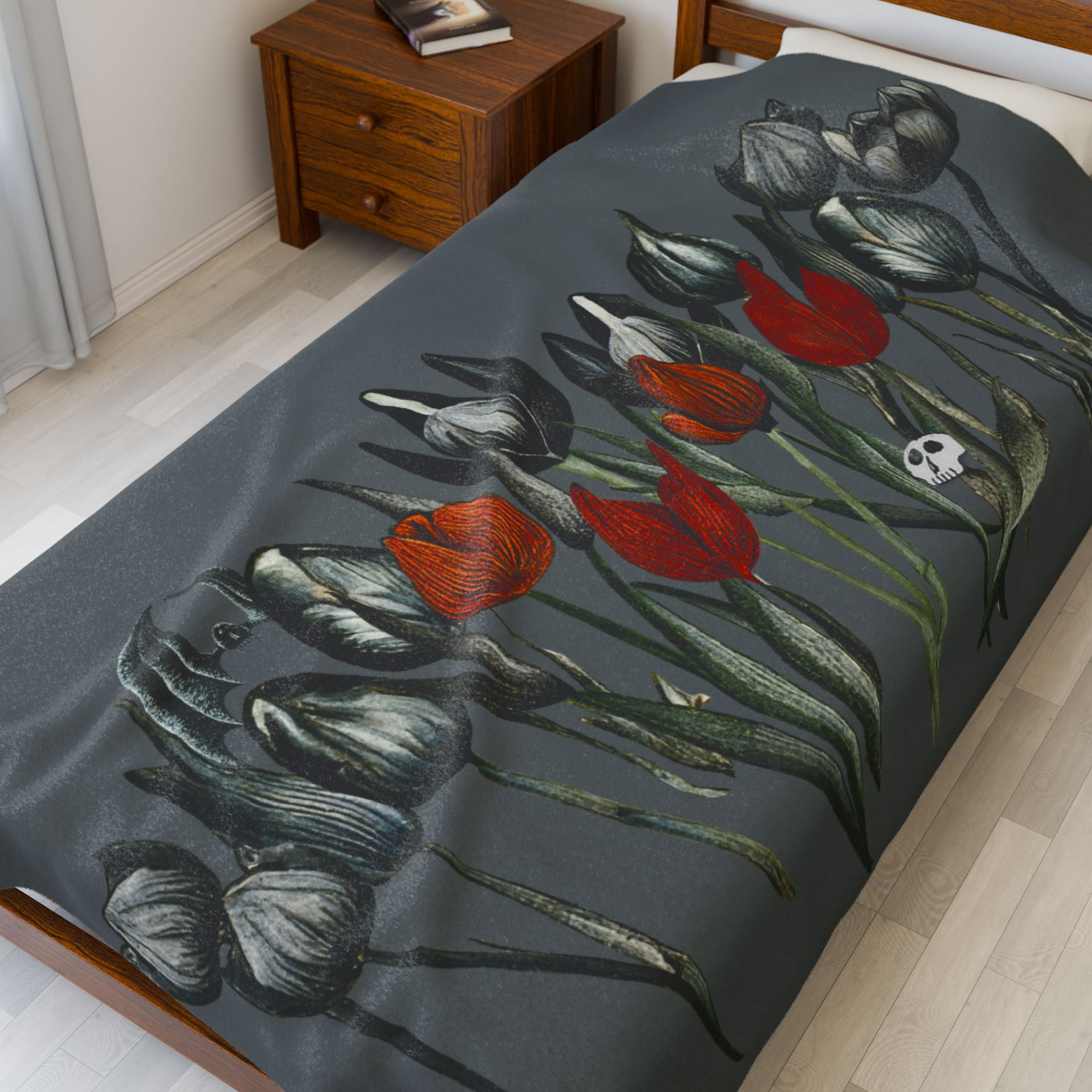 A wooden bed features a Printify Gothic Floral Velveteen Plush Blanket in dark gray with red and gray skeletal-style tulips. Beside it, a wooden nightstand holds a book on light wood flooring, creating an aura of gothic elegance.