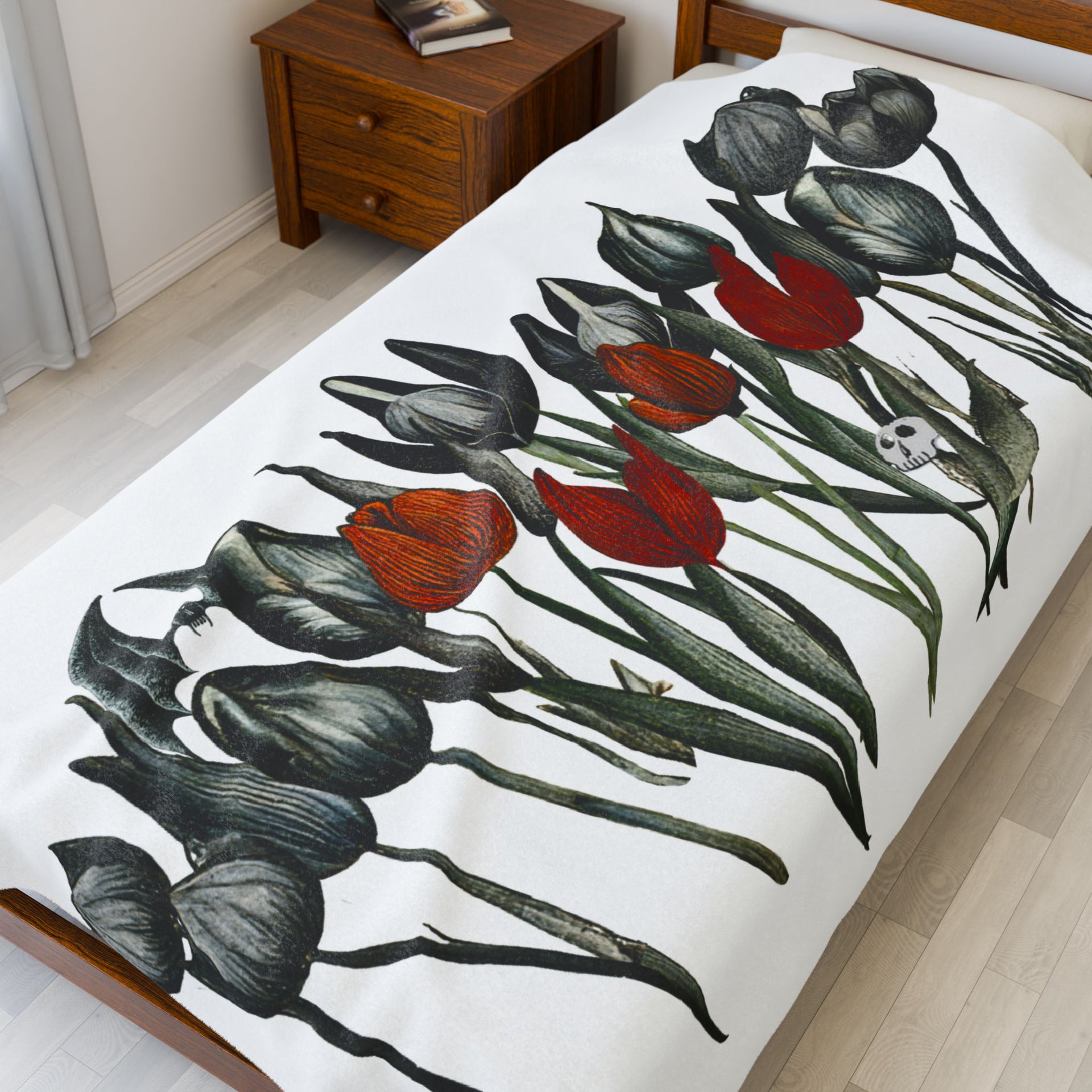 The Printify Gothic Floral Velveteen Plush Blanket in white, featuring black and red tulips, adds a touch of gothic elegance to the bed. The room is enhanced by wooden furniture like a nightstand with a book and a light wood floor.