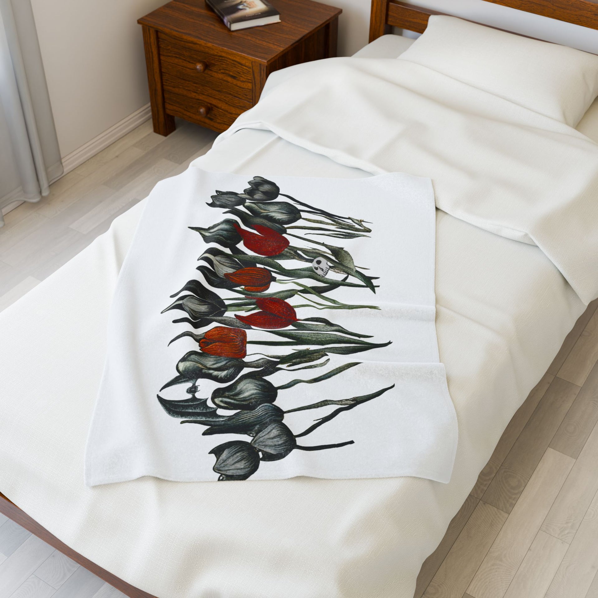 In a bright room, a white bed is paired with a wooden nightstand. The Gothic Floral Velveteen Plush Blanket – White by Printify, adorned with black and red tulips, drapes across the bed for a touch of velveteen charm.