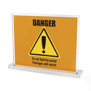 The Printify "DANGER: Do not feed the portal. Packages will vanish" acrylic sign features bold orange text and a warning triangle. Displayed in a clear stand, it’s a customizable, unique package delivery sign for your desk or entryway.