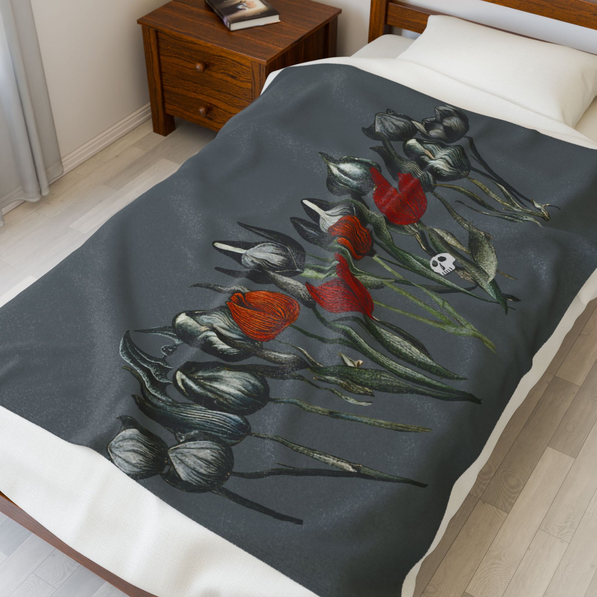 In a bright room, a bed adorned with Printifys Gothic Floral Velveteen Plush Blanket – Dark Grey, featuring red and gray floral art, exudes gothic elegance. A wooden nightstand nearby holds an intriguing book.
